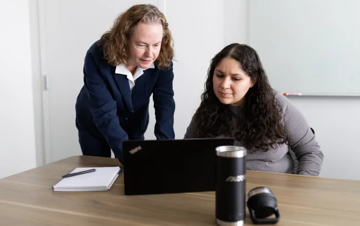 Two employees consult over a laptop at a desk.