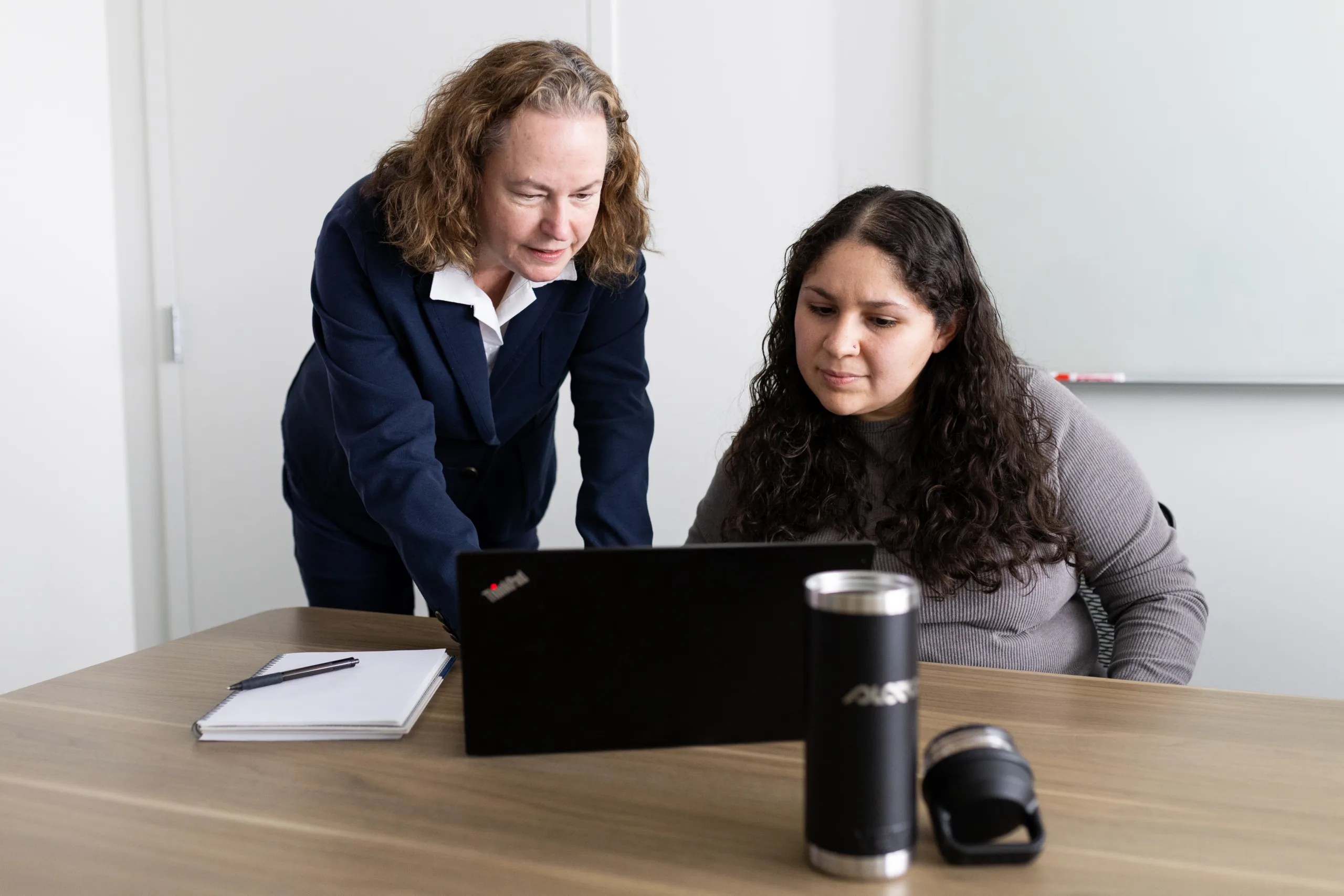 Two employees consult over a laptop at a desk.