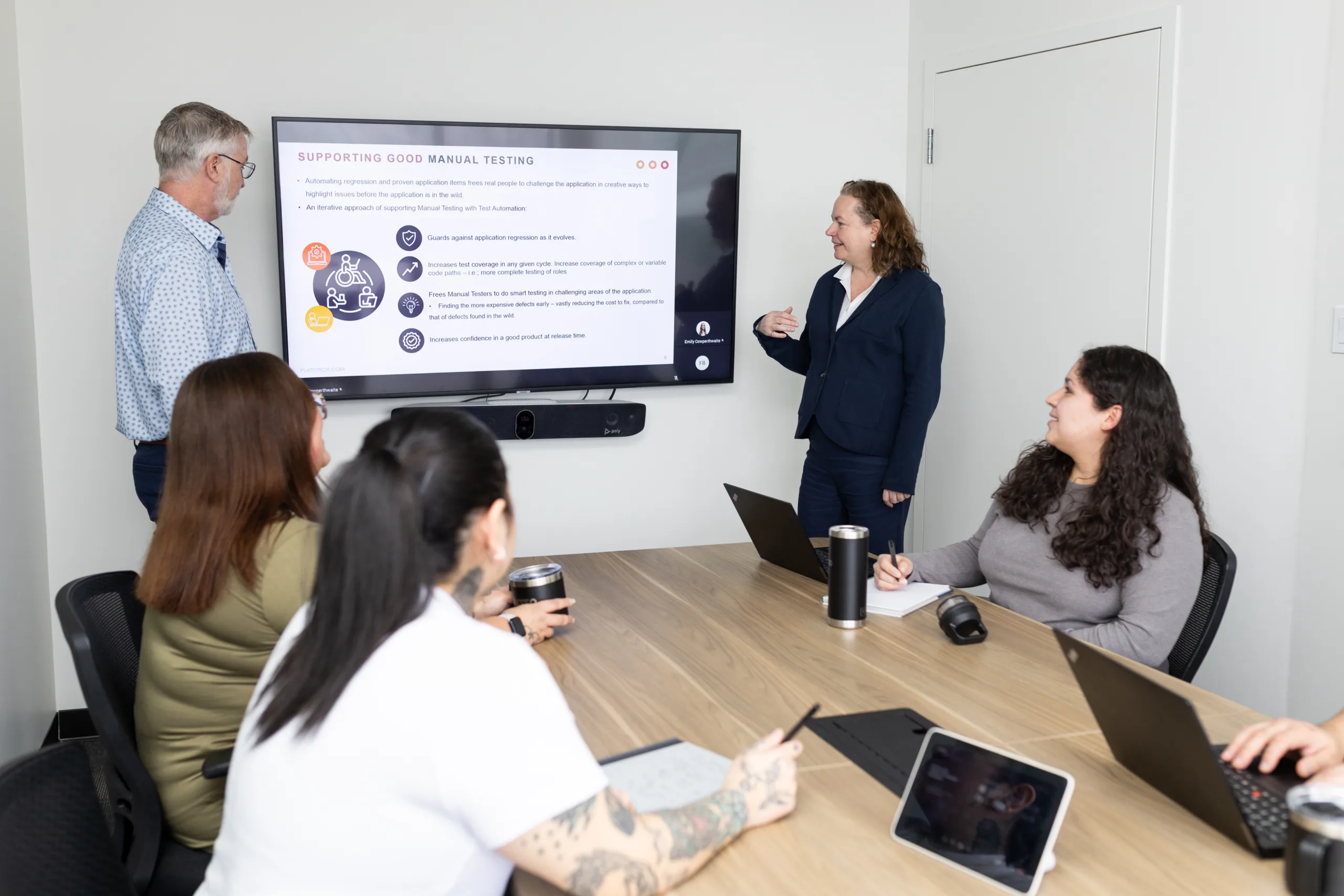 Group of people seated at a boardroom table with laptops and notebooks, while two individuals stand at the front presenting.