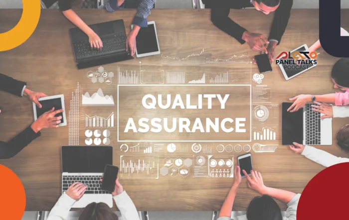 Top view of a boardroom table with multiple people sitting around on phones, tablets and laptops with graphics and icons relating to the title in the centre of the table saying Quality Assurance
