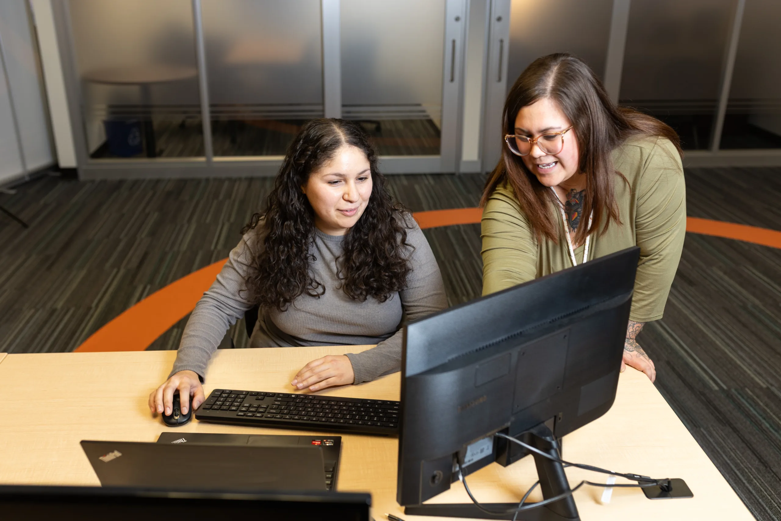 Two women collaborating in a workspace; one is seated with a desktop while the other stands and leans in to talk.