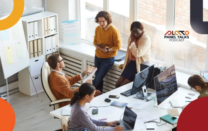 A group of coworkers in a bright modern office collaborate around desks with laptops and monitors; two people stand talking by the window while others work seated, with the PLATO Panel Talks Podcast logo displayed in the corner.